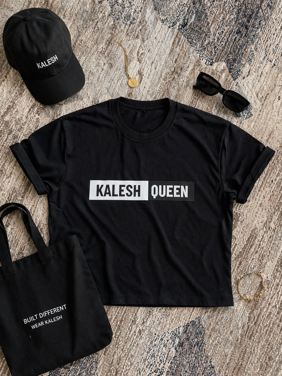 Black t-shirt with 'KALESH QUEEN' text, black cap, black tote bag, and sunglasses on a textured surface.